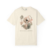 An off-white short-sleeve crewneck t-shirt featuring a watercolor floral graphic in soft pink, beige, and green tones, with the text 'ROOTED & RISING' printed below the flowers.
