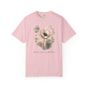 A light pink short-sleeve crewneck t-shirt featuring a watercolor-style floral graphic in the center with the text 'ROOTED & RISING' printed below it, displayed on a clean white background.