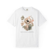 A white short-sleeve crewneck t-shirt featuring a watercolor floral graphic in soft pink, beige, and green tones, with the text 'ROOTED & RISING' printed below the flowers.