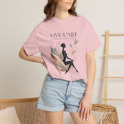 A woman wearing a light pink graphic t-shirt with the French text 'VIVE L'ART' and a black silhouette of a seated woman surrounded by leaves, paired with light blue denim shorts in a warm indoor setting.