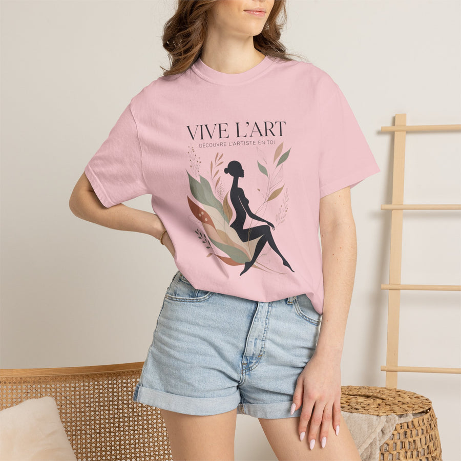 A woman wearing a light pink graphic t-shirt with the French text 'VIVE L'ART' and a black silhouette of a seated woman surrounded by leaves, paired with light blue denim shorts in a warm indoor setting.