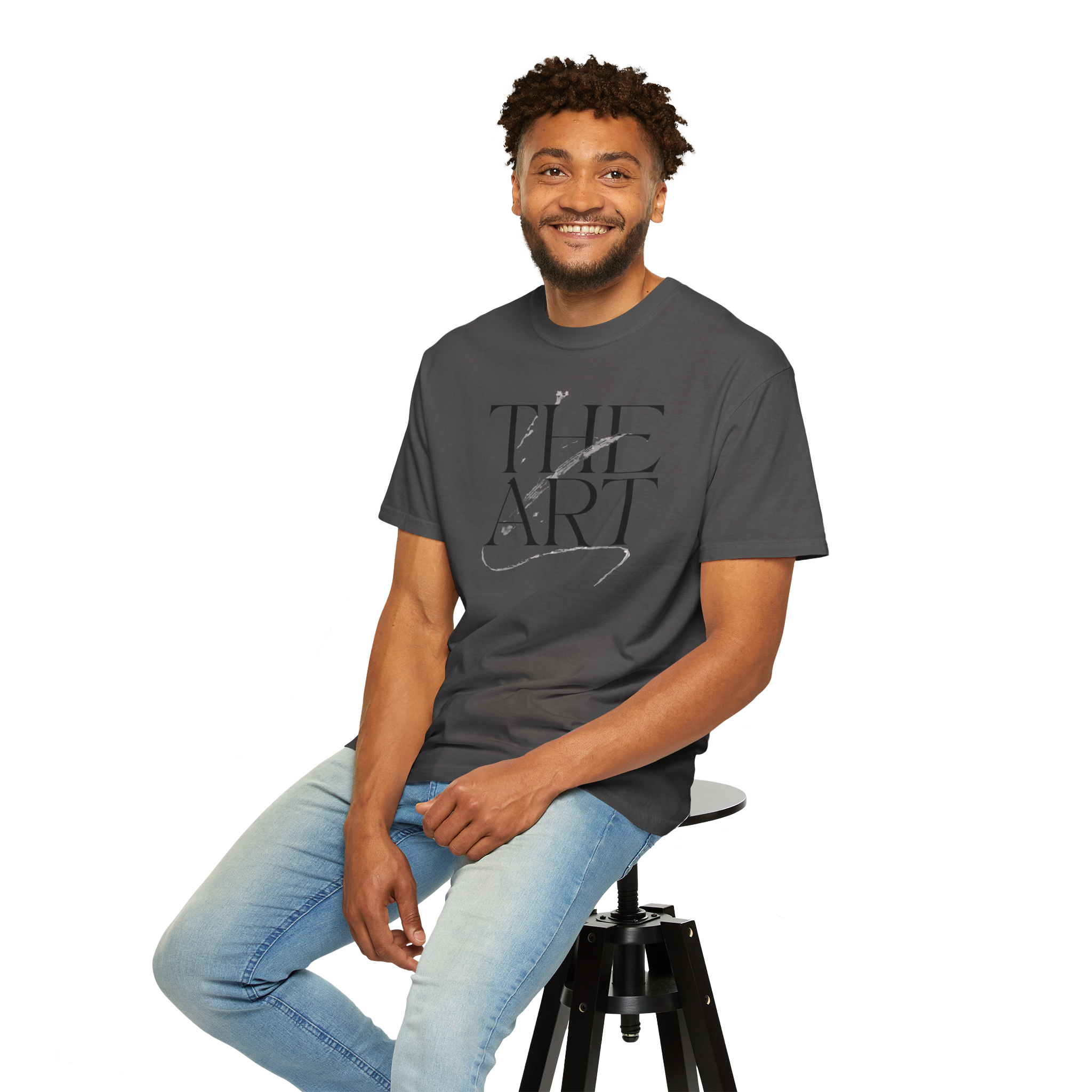 A man sitting on a black stool wearing a dark gray t-shirt with a black graphic print that reads 'THE ART' and light blue jeans, against a plain white background.